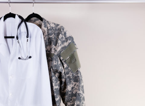 Medical Doctor Consultation white coat, stethoscope around collar, and military uniform hanging on rack. Off white wall in background. Horizontal layout with copy space.